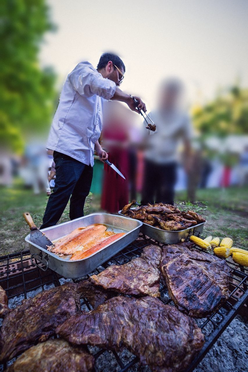 Chef sirviendo carnes, salmón y choclos en la parrilla, con invitados desenfocados al fondo
