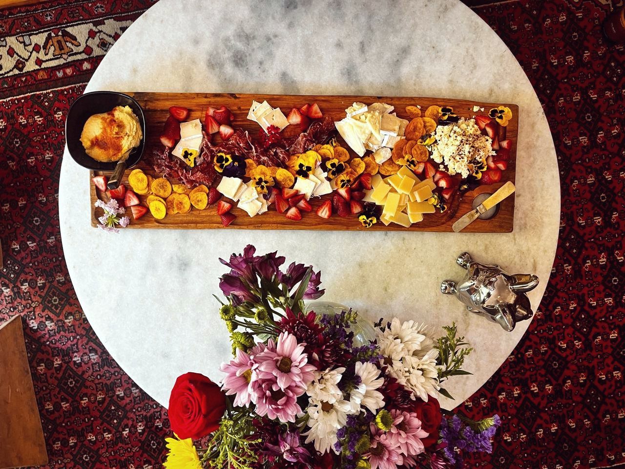Tabla de quesos, frutillas y flores comestibles sobre mármol, con un ramo de flores de estación
